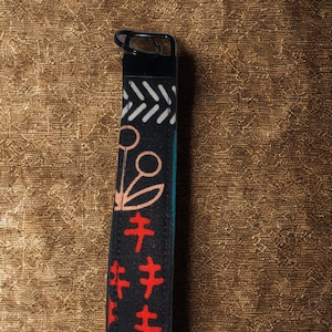 Könnte beinhalten: Schwarzer Stoff-Schlüsselanhänger mit Metallring. Der Schlüsselanhänger hat ein geometrisches Muster in Weiß, Türkis und Rot. Das Design umfasst florale und abstrakte Formen. Der Hintergrund ist ein strukturiertes Braun.