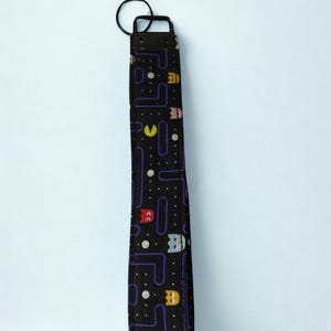 May include: Black wrist strap with a Pac-Man pattern. The strap features a repeating design of Pac-Man characters, ghosts, and maze elements in various colors. A black metal ring is attached at the top.