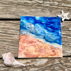 May include: A square resin art piece depicting a beach scene with blue water, white foam, and a sandy beach. The piece is on a wooden surface with a seashell and a starfish.