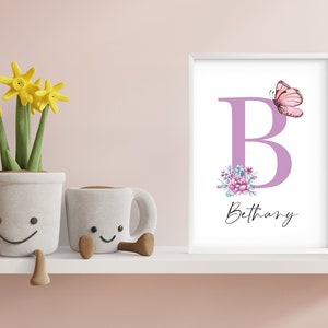 May include: A white frame with a purple letter B, pink butterfly, and floral design. The name Bethany is written in cursive below the letter. The frame is on a white shelf with three potted plants.