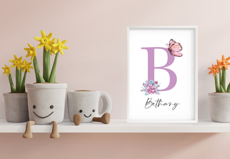 May include: A white frame with a purple letter B, pink butterfly, and floral design. The name Bethany is written in cursive below the letter. The frame is on a white shelf with three potted plants.