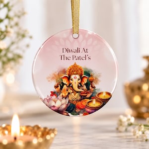 May include: A round, glossy ceramic ornament featuring a gold ribbon hanger. The design displays a colorful illustration of the deity Ganesha, a lotus flower, and two lit oil lamps. The text "Diwali At The Patel's" is printed in dark brown.