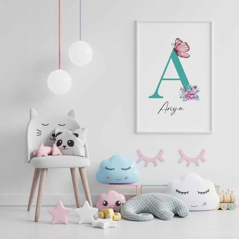 May include: A white framed print featuring a teal letter A adorned with pink flowers and a butterfly. The name Ariya is written beneath the letter. The print is displayed on a white wall in a nursery, alongside a grey cat-shaped chair, a blue cloud-shaped cushion, and other toys.