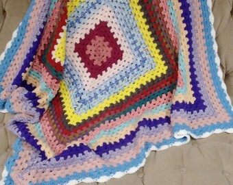 May include: A colorful, handmade crochet blanket with a square pattern. The blanket features a central maroon square, surrounded by rings of yellow, red, green, pink, and blue. The edges are finished with a white scalloped border. The blanket is draped over a beige sofa.