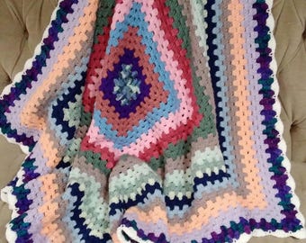 May include: A handmade, colorful crochet blanket with a square pattern. The blanket features a variety of pastel colors, including pink, blue, green, and purple, arranged in concentric squares. The edges are finished with a white, scalloped border.
