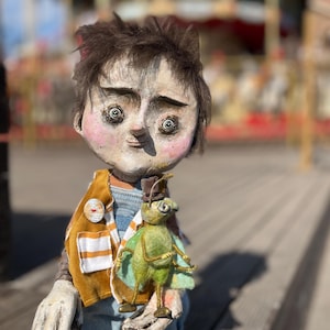 May include: A handcrafted doll with a whimsical design, wearing a mustard yellow vest over denim overalls. The doll holds a green insect figure. The doll has a unique, expressive face with large eyes and a slightly upturned nose. The background is blurred, suggesting an outdoor setting.