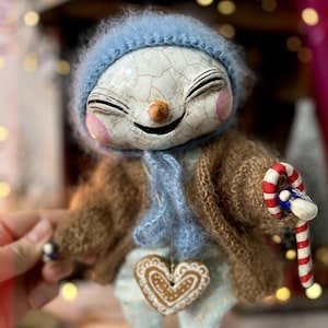 May include: A smiling snowman doll wearing a blue knitted hat and scarf, a brown sweater and holding a candy cane. The doll has a heart-shaped gingerbread cookie hanging from its neck.