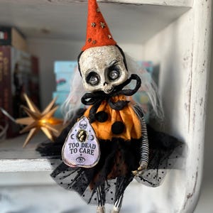 Könnte beinhalten: Eine Puppe mit rissigem, schädelartigem Kopf, orangefarbenem Kegelhut mit goldenen Sternen und schwarz-orangefarbenem Rüschenoutfit. Sie hat weißes Haar, schwarz-weiß gestreifte Gliedmaßen und einen Planchette-Anhänger mit der Aufschrift TOO DEAD TO CARE.