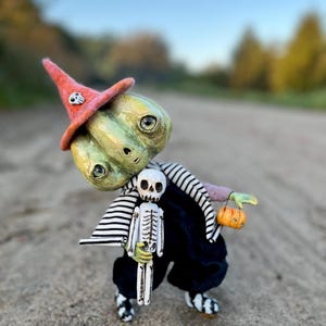 Może przedstawiać: Kapryśna figurka na Halloween przedstawiająca postać o zielonej skórze w czerwonym kapeluszu czarownicy z czaszką, czarnych spodniach i pasiastą koszulą. Trzyma mały szkielet i dynię. Figurka jest ustawiona na teksturowanej powierzchni.