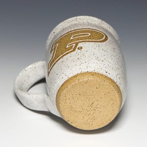 May include: A speckled white ceramic mug with a large handle. The mug features a gold-colored letter "P" with a white outline. The bottom of the mug is a light brown color with a speckled texture and a handwritten signature.