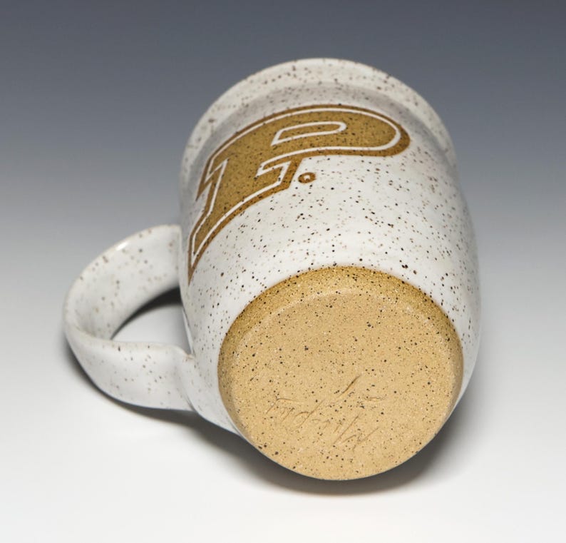 May include: A speckled white ceramic mug with a large handle. The mug features a gold-colored letter "P" with a white outline. The bottom of the mug is a light brown color with a speckled texture and a handwritten signature.