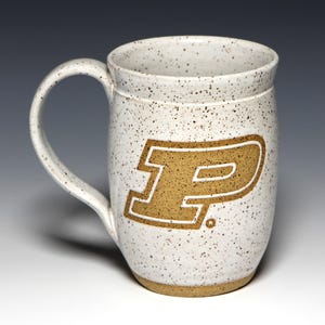 May include: A speckled white ceramic mug with a large handle. The mug features a gold-colored letter "P" with a white outline. The mug has a wide, cylindrical shape and a light brown base.