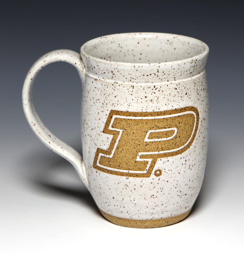 May include: A speckled white ceramic mug with a large handle. The mug features a gold-colored letter "P" with a white outline. The mug has a wide, cylindrical shape and a light brown base.