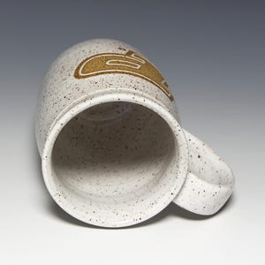 May include: A speckled ceramic mug with a curved handle. The mug is off-white with brown speckles and a gold-colored design on the side. The mug is empty and angled towards the viewer.