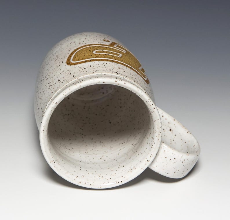 May include: A speckled ceramic mug with a curved handle. The mug is off-white with brown speckles and a gold-colored design on the side. The mug is empty and angled towards the viewer.