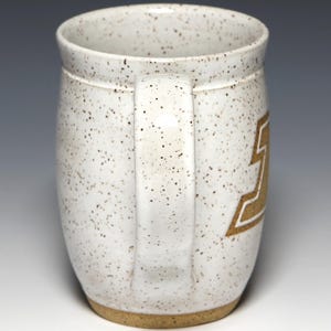 May include: A speckled white ceramic mug with a brown handle and base. The mug features a gold-colored design on the side. The mug is designed for hot beverages.