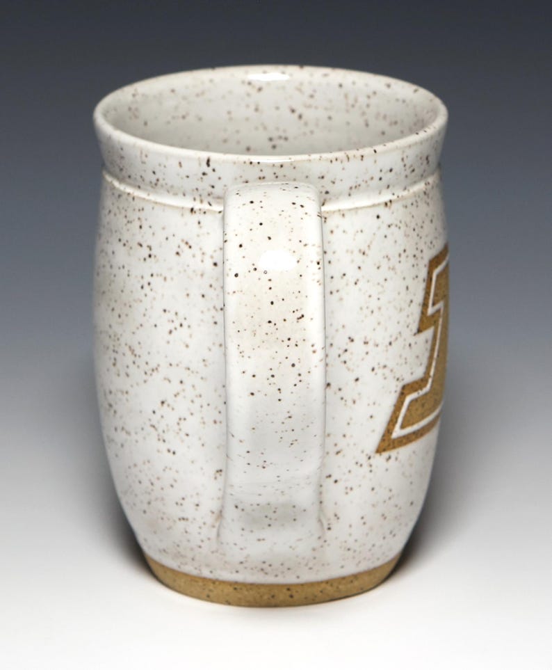 May include: A speckled white ceramic mug with a brown handle and base. The mug features a gold-colored design on the side. The mug is designed for hot beverages.