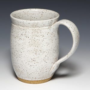May include: A speckled white ceramic mug with a large handle. The mug has a cylindrical shape with a slightly flared rim and a tan base. The white surface is covered in small, dark brown speckles.