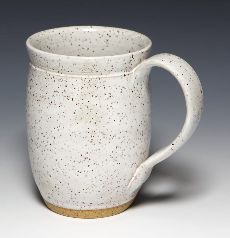 May include: A speckled white ceramic mug with a large handle. The mug has a cylindrical shape with a slightly flared rim and a tan base. The white surface is covered in small, dark brown speckles.