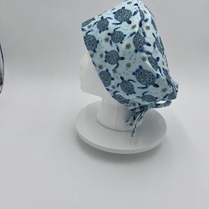 May include: A light blue surgical scrub hat with a pattern of blue and green sea turtles. The hat has a tie at the back.