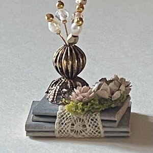 May include: A stack of three grey books with a lace trim and a decorative vase with a floral arrangement on top. The vase is gold and has a beaded stem with white and gold beads.