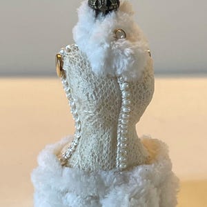 May include: A cream-coloured dress form ornament with a lace-like texture, faux pearl detailing, and fluffy white trim. It is topped with a small, ornate crown. The ornament is designed to resemble a dress.