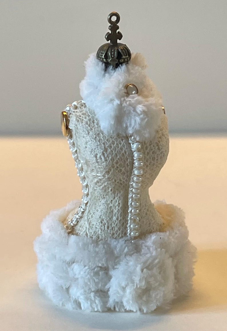 May include: A cream-coloured dress form ornament with a lace-like texture, faux pearl detailing, and fluffy white trim. It is topped with a small, ornate crown. The ornament is designed to resemble a dress.