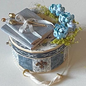 May include: A round, blue and white patterned box with a gold trim and a fleur-de-lis design. Two small grey books are stacked on top of the box, tied with a white ribbon and decorated with gold pins. A cluster of light blue flowers with green leaves sits on top of the books.
