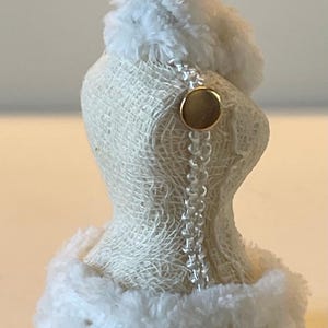 May include: A miniature dress form ornament with a cream-coloured, woven fabric body. The form is trimmed with fluffy white faux fur at the top and bottom. A gold-coloured button and a string of clear beads adorn the back. A decorative metal cap tops the form.