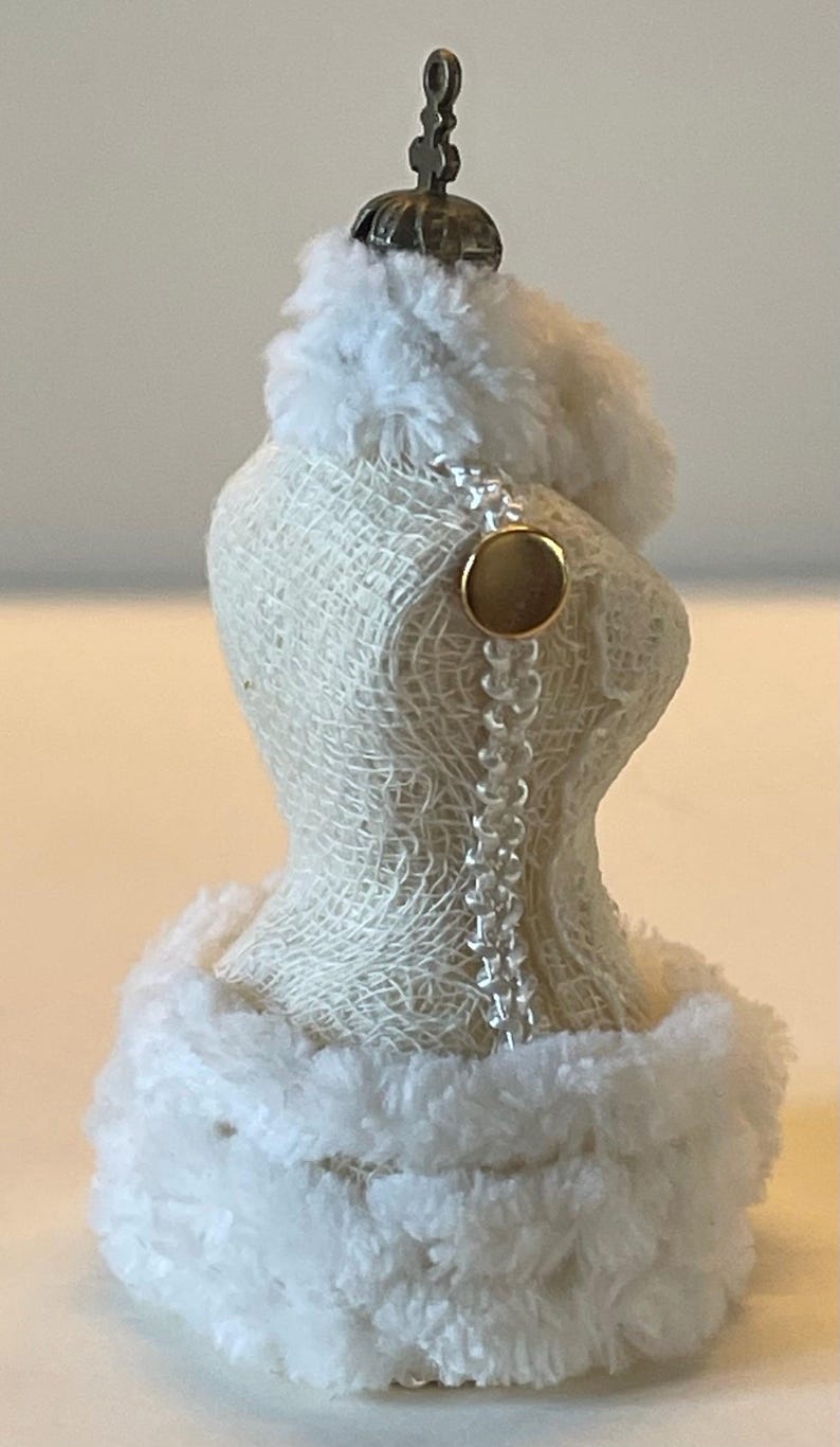 May include: A miniature dress form ornament with a cream-coloured, woven fabric body. The form is trimmed with fluffy white faux fur at the top and bottom. A gold-coloured button and a string of clear beads adorn the back. A decorative metal cap tops the form.
