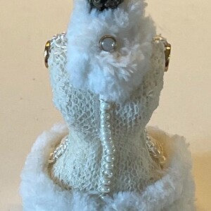 May include: A miniature white dress form with a decorative crown and pearl accents. The dress form is covered in a textured, open-weave fabric and trimmed with fluffy white material. A string of pearls runs down the back.