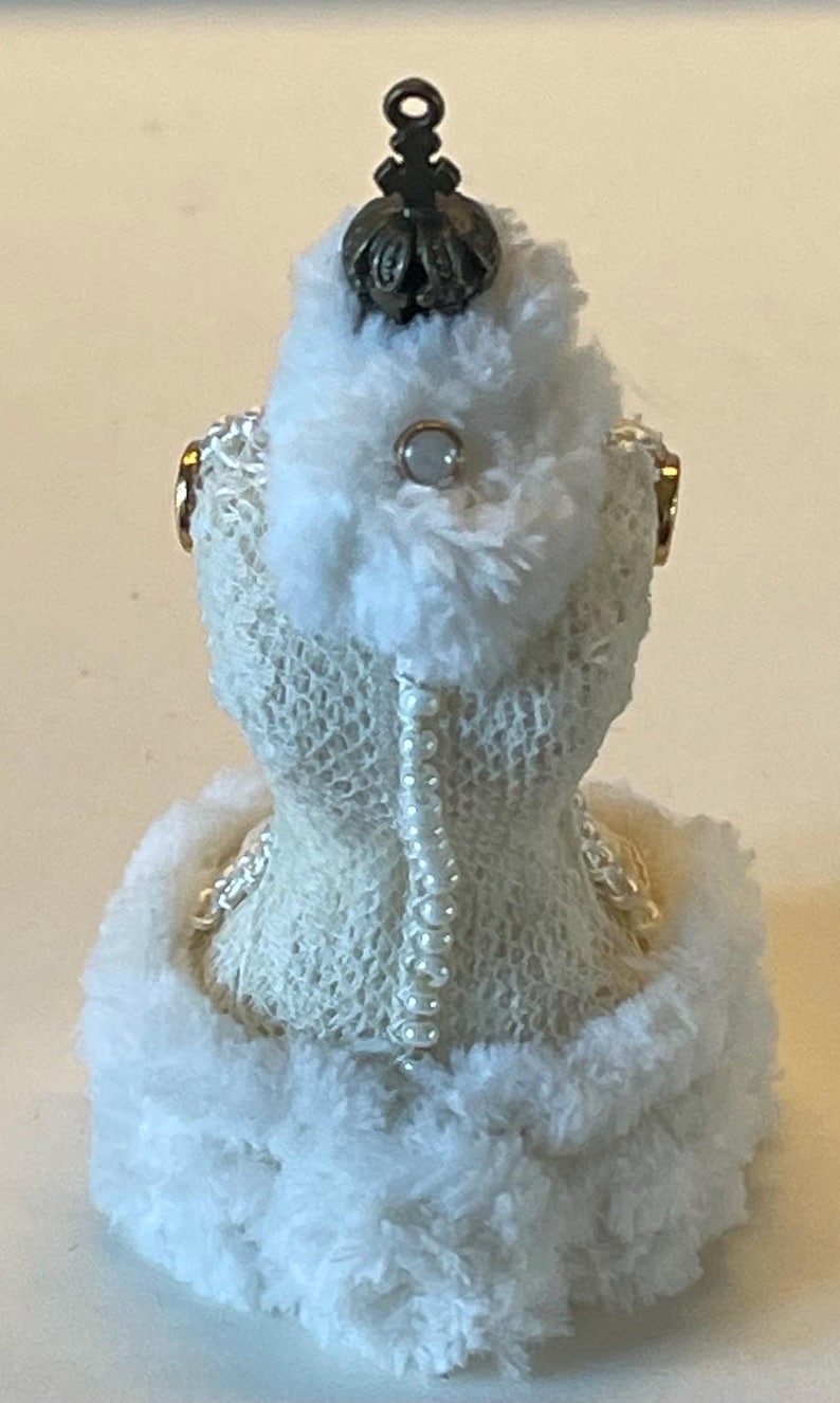 May include: A miniature white dress form with a decorative crown and pearl accents. The dress form is covered in a textured, open-weave fabric and trimmed with fluffy white material. A string of pearls runs down the back.
