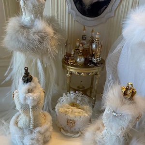May include: A miniature vanity scene in white and gold, featuring a portrait, perfume bottles, and decorative dress forms. The dress forms are decorated with lace, feathers, and faux fur. A small gold table holds perfume bottles with pearl details.