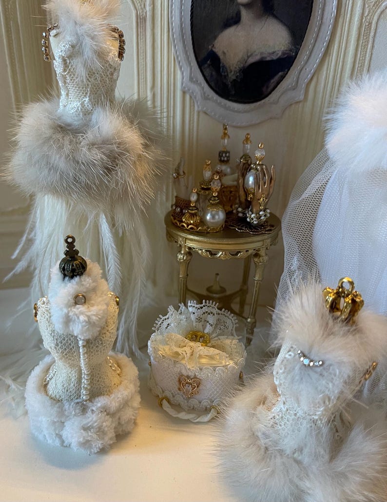 May include: A miniature vanity scene in white and gold, featuring a portrait, perfume bottles, and decorative dress forms. The dress forms are decorated with lace, feathers, and faux fur. A small gold table holds perfume bottles with pearl details.