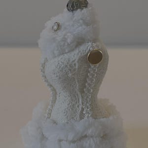 May include: A white Christmas tree decoration in the shape of a dress, featuring a fluffy faux fur trim and details. The dress is made of a delicate, open-weave fabric and decorated with a string of pearls. A small, metallic cap tops the ornament.