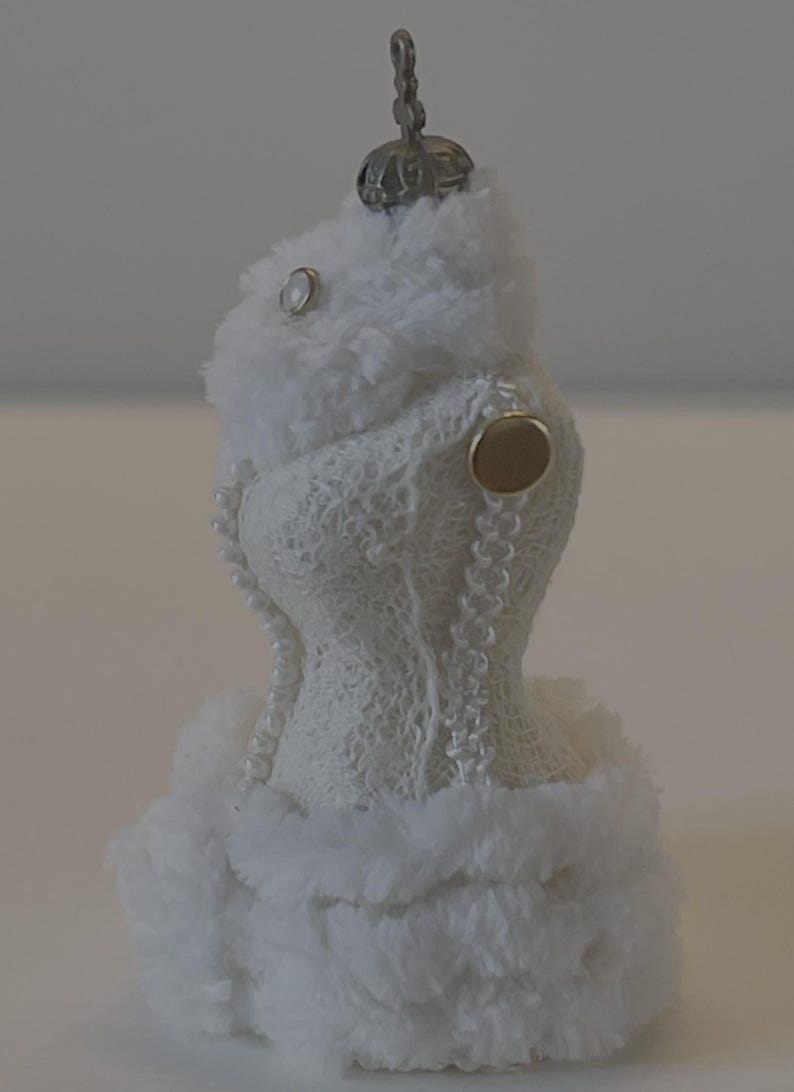 May include: A white Christmas tree decoration in the shape of a dress, featuring a fluffy faux fur trim and details. The dress is made of a delicate, open-weave fabric and decorated with a string of pearls. A small, metallic cap tops the ornament.