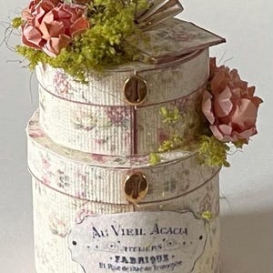 May include: Two round, white boxes with floral patterns and gold accents. The boxes are stacked on top of each other and decorated with pink flowers and green moss. The bottom box has a label that reads "Au Viel Acacia 1771 Paris - Fabrique 21 Rue de la Vieille Touque".