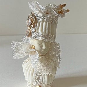 May include: A white ceramic doll head with a white lace and fabric hat with gold accents. The doll is wearing a white lace collar.
