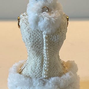 May include: A miniature dress form ornament, featuring a cream-coloured, textured bodice with a row of pearls down the back. The dress form is trimmed with fluffy white faux fur and topped with a decorative bronze crown.