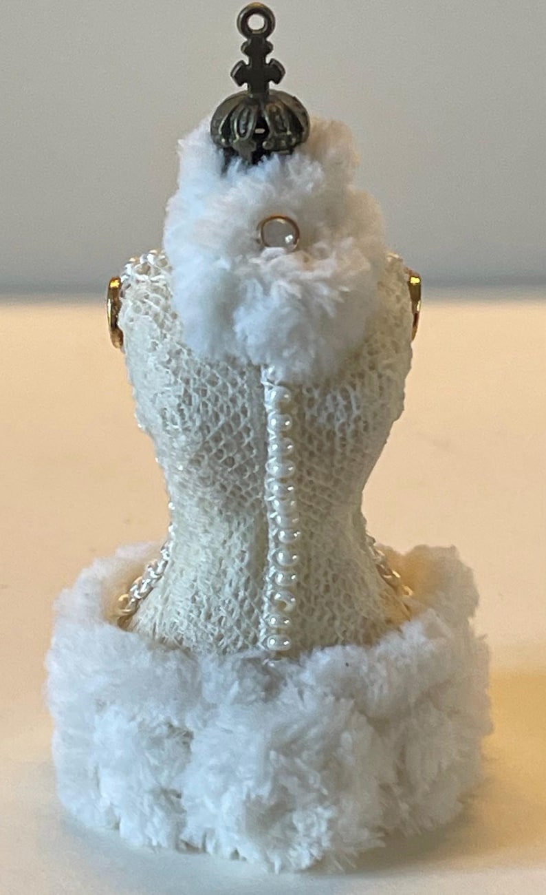 May include: A miniature dress form ornament, featuring a cream-coloured, textured bodice with a row of pearls down the back. The dress form is trimmed with fluffy white faux fur and topped with a decorative bronze crown.