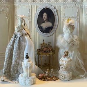 May include: A detailed miniature scene with doll dresses, a portrait, and perfume bottles. The dresses are cream and white, with lace and feather embellishments. A small gold table and tray display the perfume bottles and floral arrangements.