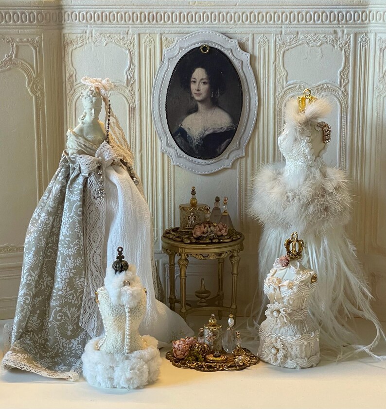 May include: A detailed miniature scene with doll dresses, a portrait, and perfume bottles. The dresses are cream and white, with lace and feather embellishments. A small gold table and tray display the perfume bottles and floral arrangements.