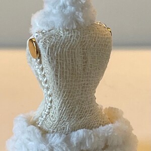 May include: A miniature white dress form with a textured, off-white fabric body. The dress form is trimmed with fluffy white faux fur at the top and bottom. Gold-coloured accents and a decorative metal crown add detail.