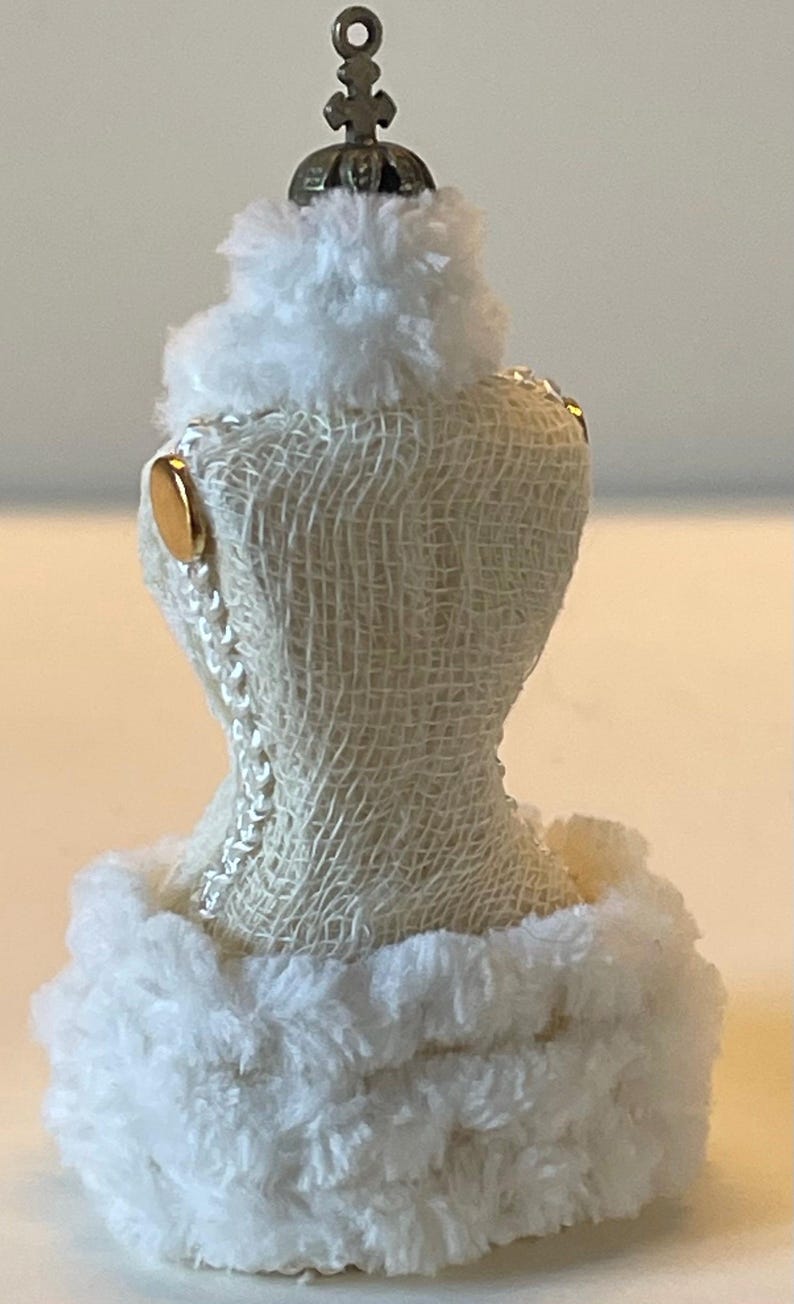 May include: A miniature white dress form with a textured, off-white fabric body. The dress form is trimmed with fluffy white faux fur at the top and bottom. Gold-coloured accents and a decorative metal crown add detail.