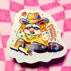 May include: A holographic sticker featuring a raccoon wearing a colorful cowboy hat, a yellow vest, and a bandana. The raccoon has a red nose and is holding its head with both hands. The word PANIC is printed in rainbow letters on the right side.