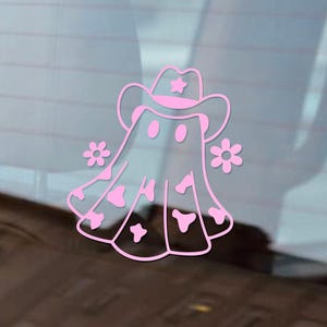 May include: A pink vinyl sticker featuring a cartoon ghost wearing a cowboy hat with a star and two flowers. The ghost has a cow-print design on its body. The sticker is designed to be applied to a car window or other smooth surface.