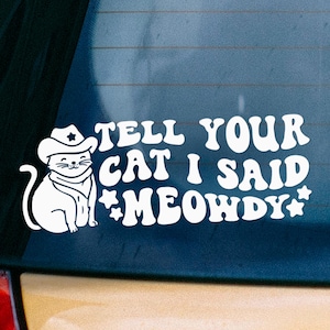 May include: A white vinyl decal applied to a car window featuring a graphic of a cat wearing a cowboy hat and bandana. The text reads, "TELL YOUR CAT I SAID MEOWDY," accompanied by small star icons on either side of the final word.