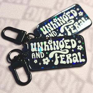 May include: Two rectangular keychains with the words "UNHINGED AND FERAL" in white, retro-style font. The keychains are dark blue with holographic glitter and small flower and star accents. Each keychain has a black metal clasp.
