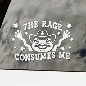 May include: White vinyl decal featuring a cartoon frog wearing a cowboy hat with the text "THE RAGE CONSUMES ME". The frog has its arms raised and is surrounded by stars. The decal is applied to a car window.