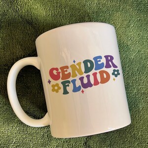 May include: White ceramic mug with a rainbow-colored design that says "GENDER FLUID" with flower and diamond accents.